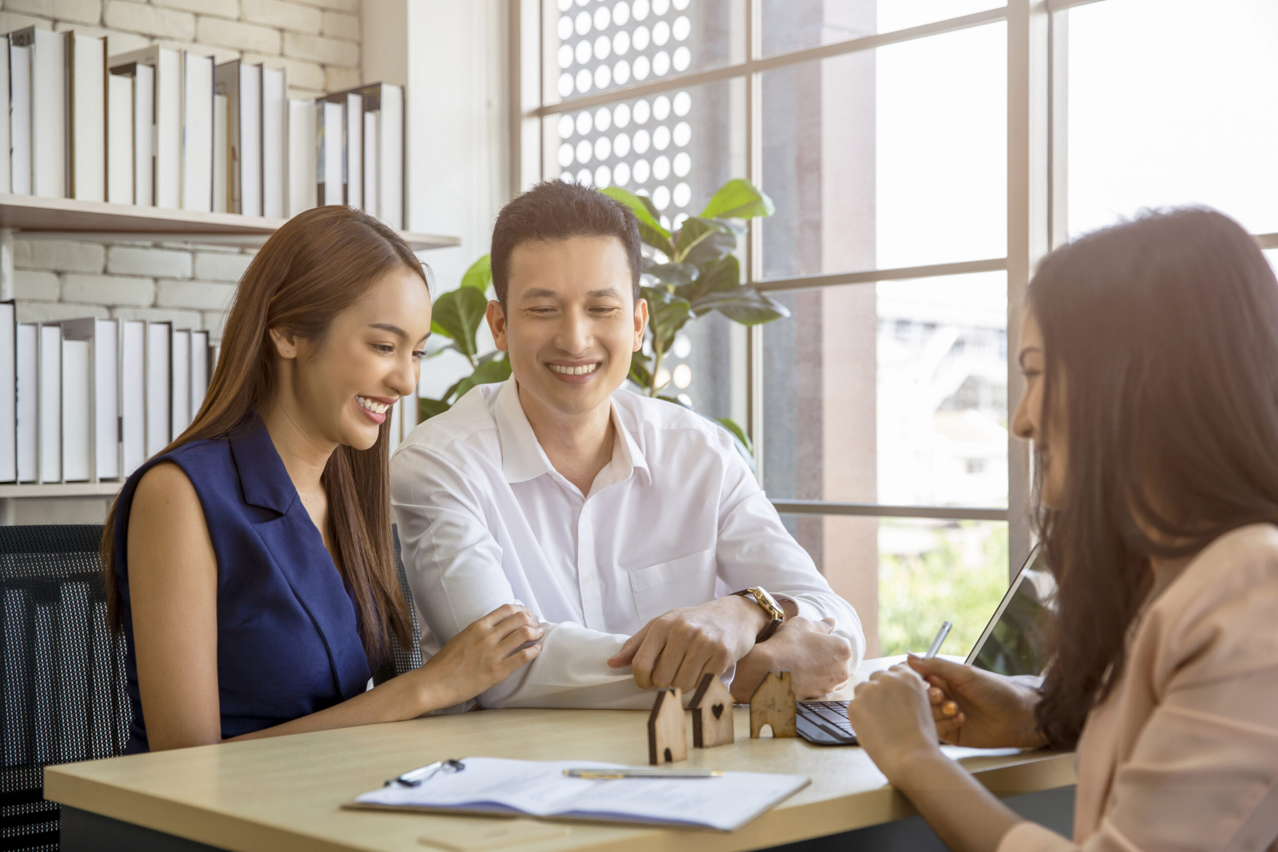 A couple smiles while discussing plans with a real estate agent at a desk, with a model house and paperwork in view.